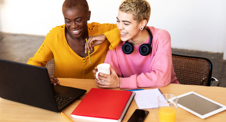 person in yellow sweater leaning on person in pink sweater looking at laptop