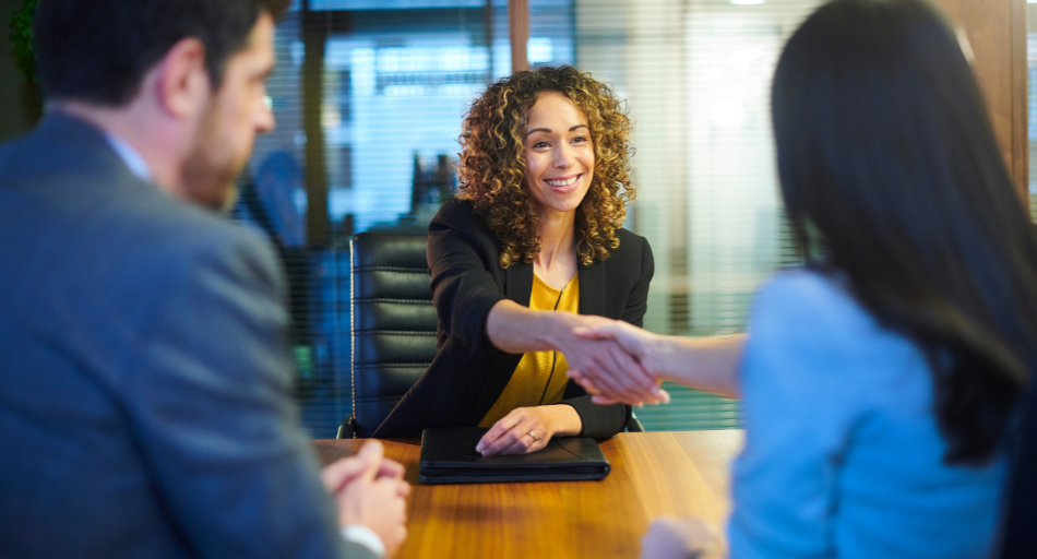 woman wearing blazer shaking hands with woman sitting across table from her