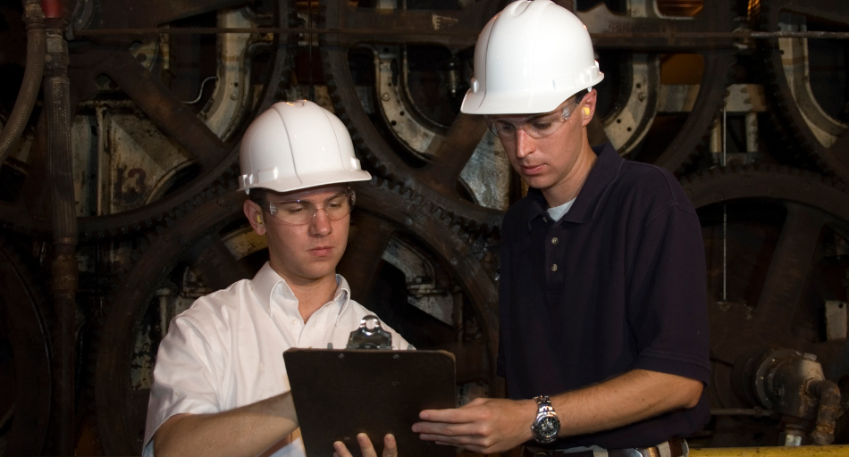 two men looking at clipboard, wearing white hard hats standing in front of industrial background