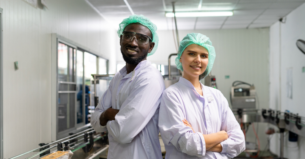 two smiling professionals in food manufacturing plant wearing hairnets and uniforms standing back-to-back with arms folded