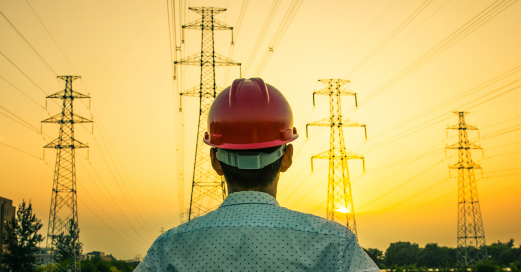 electrical engineer wearing red hard hat facing away from camera, looking toward transmission towers on sunset horizon