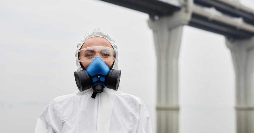 Person wearing full HAZMAT PPE in front of grey skies with tall concrete overpass infrastructure in the background