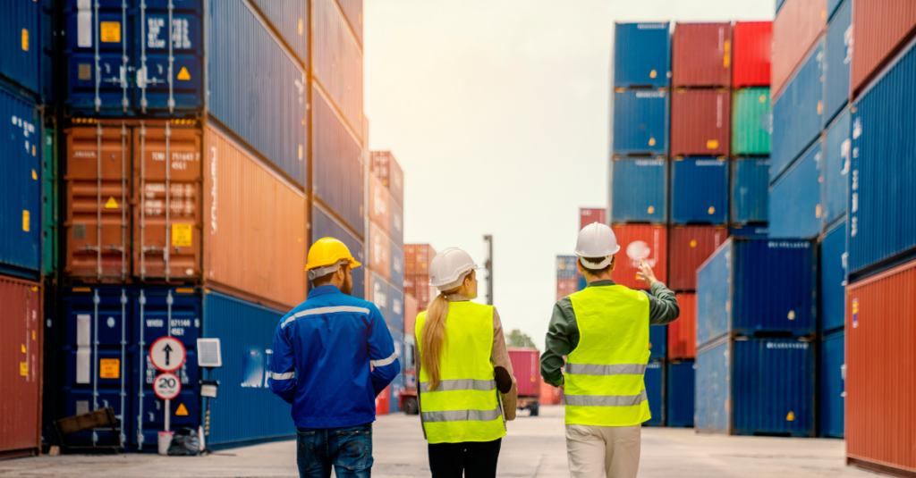 three supply chain workers in hi viz vests and hard hats walking amongst shipping containers